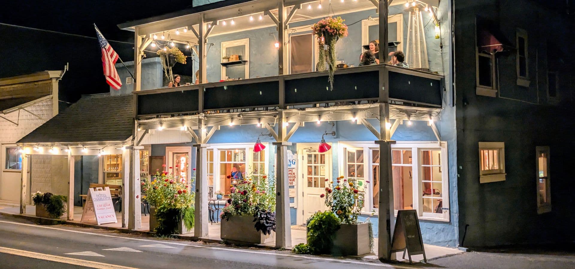A charming blue building with string lights and an American flag, featuring outdoor seating surrounded by flowers at night.