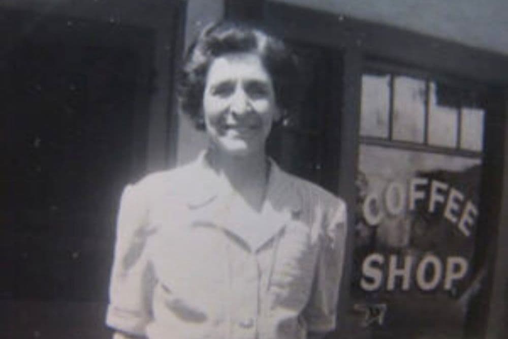 A smiling woman stands in front of a coffee shop.