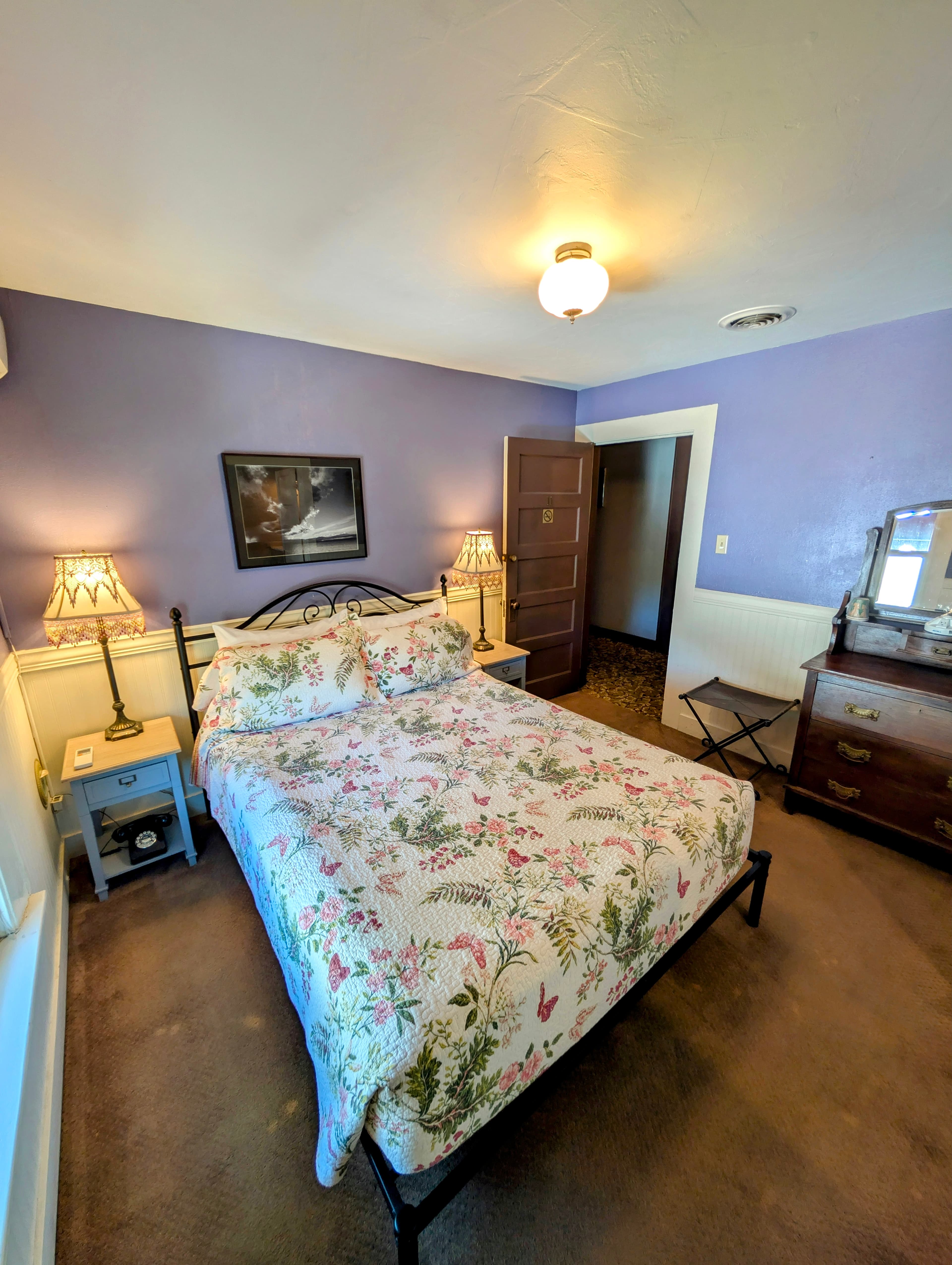 A bright and welcoming room with lavender walls, a queen bed covered with a floral quilt, and white wainscoting.