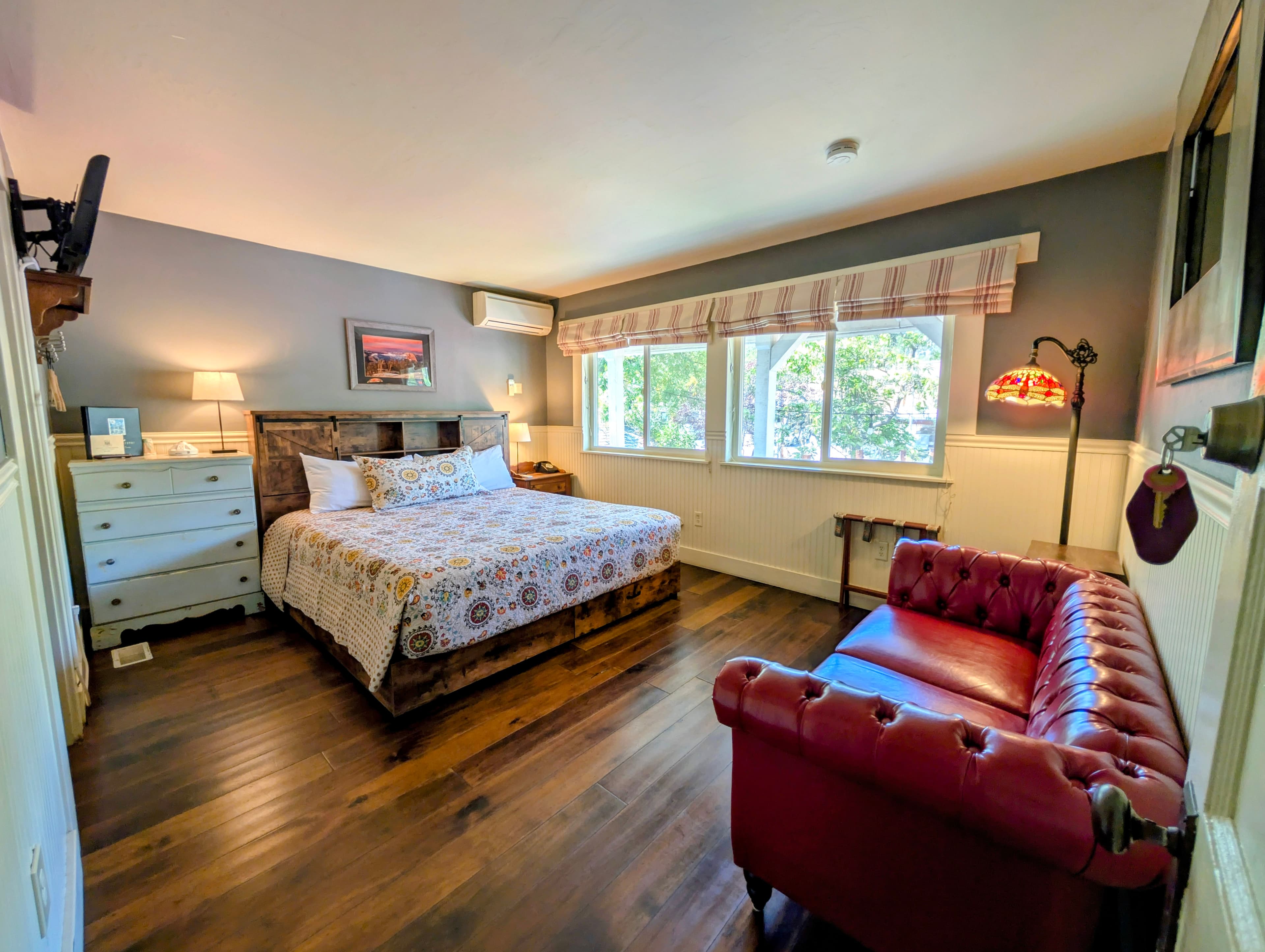 A spacious hotel room with a wooden floor, a king bed with a floral quilt, and a red couch. There's a TV mounted on the wall, and windows allow for plenty of natural light.