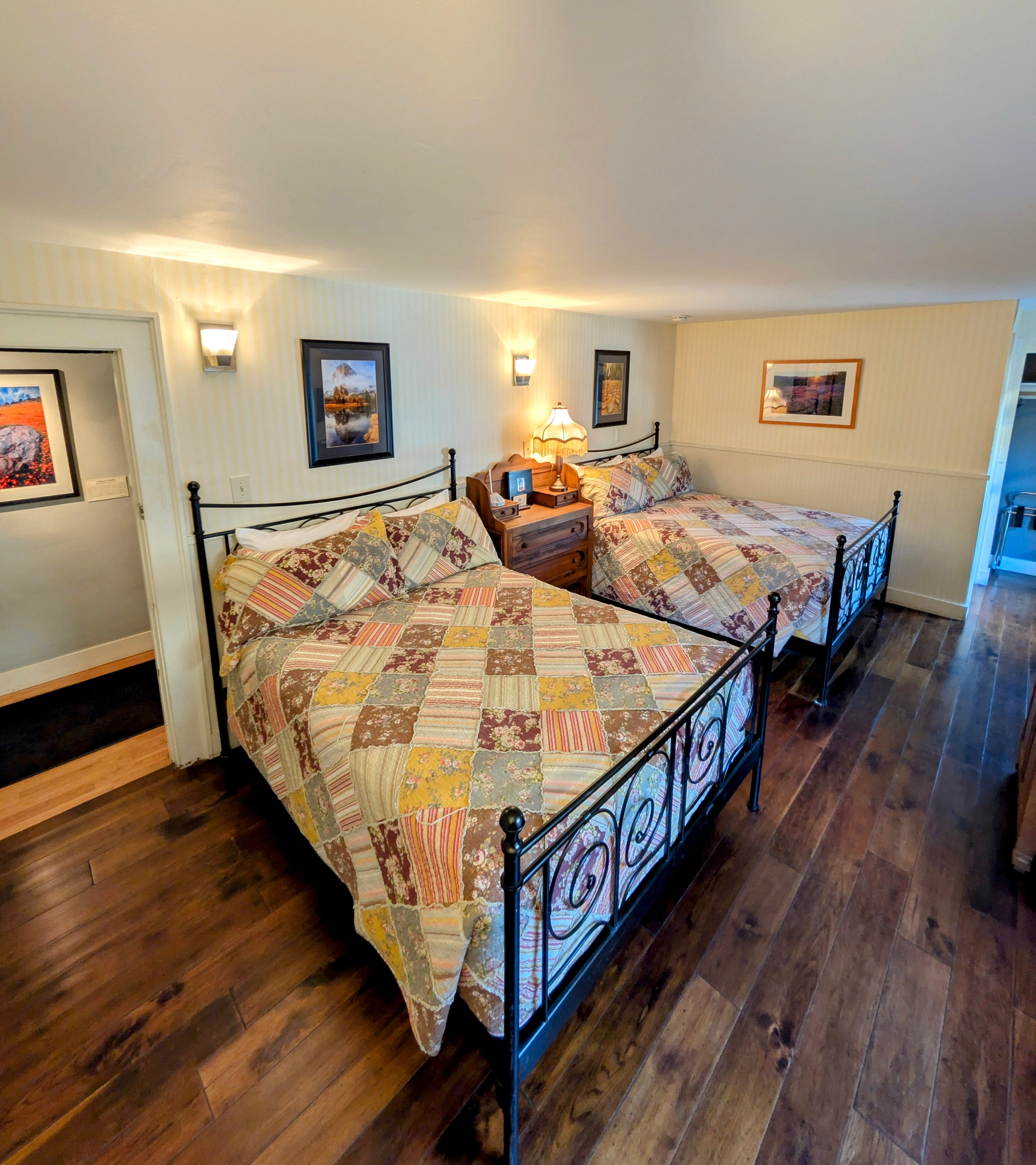 A cozy hotel room featuring two queen-size beds with colorful, patchwork quilts. The room has hardwood floors, light walls, and warm lighting.